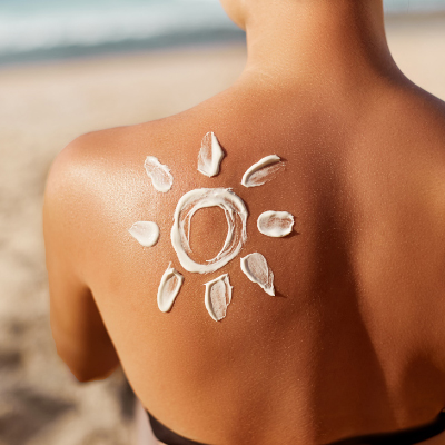 A woman is on a beach. The image shows her back only, and there is sunscreen on her left shoulder in the shape of a sun.