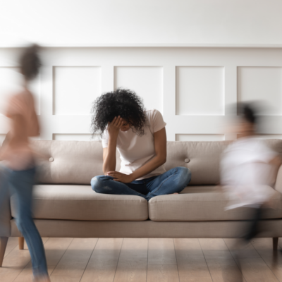 Woman sitting down on a couch with her head in her hand. Children are running around her with a motion blur effect. 