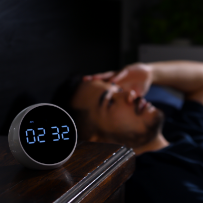 An alarm clock showing the time 2:32 AM. In the background, a young man is lying in bed awake, with an exasperated expression on his face.