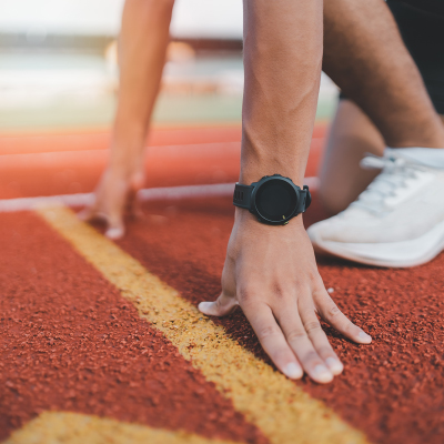 A close-up of a man’s hands and feet on a track oval as he is getting ready to start running. There is a black sports watch clearly visible on the man’s wrist. 
