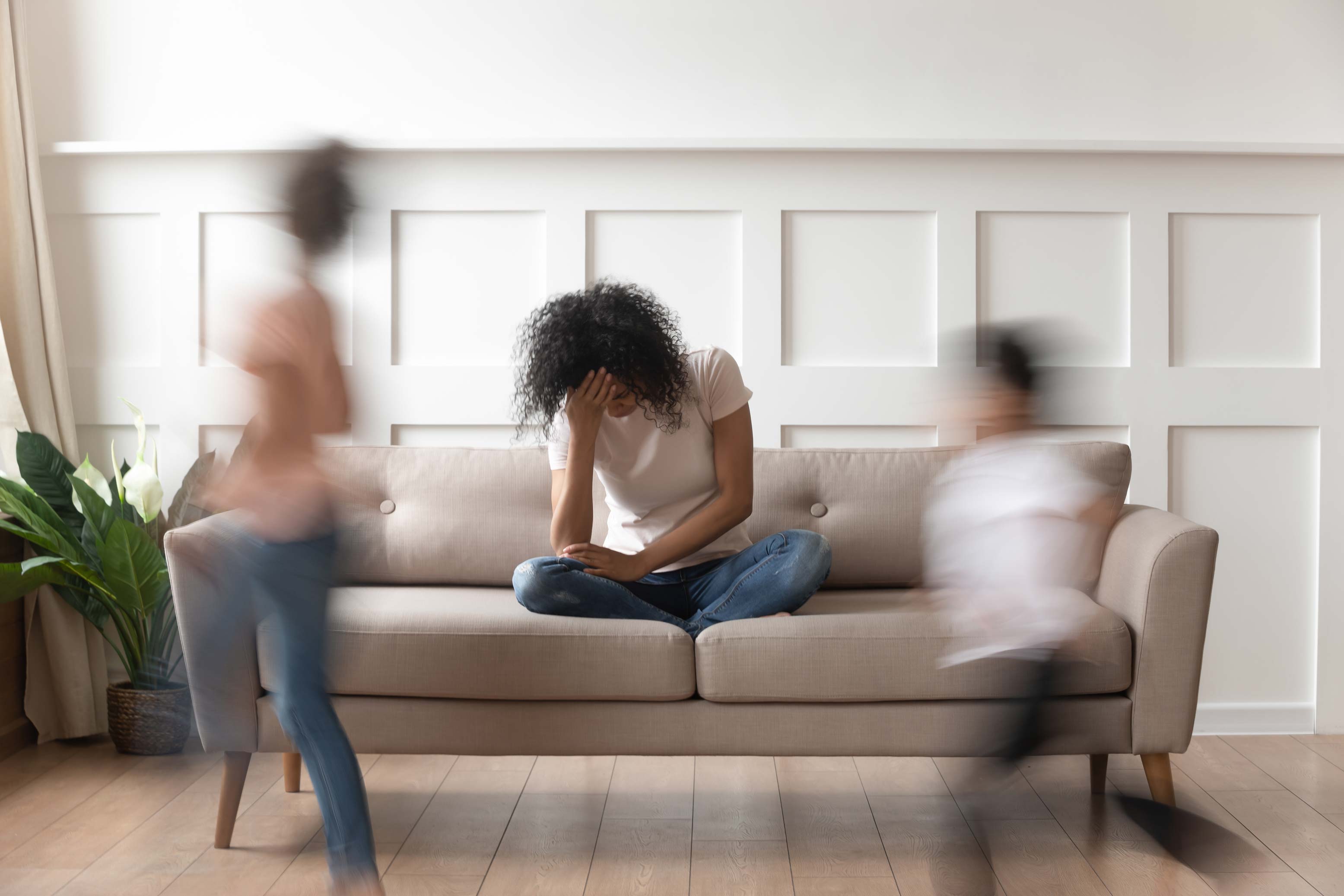 Woman sitting down on a couch with her head in her hand. Children are running around her with a motion blur effect.