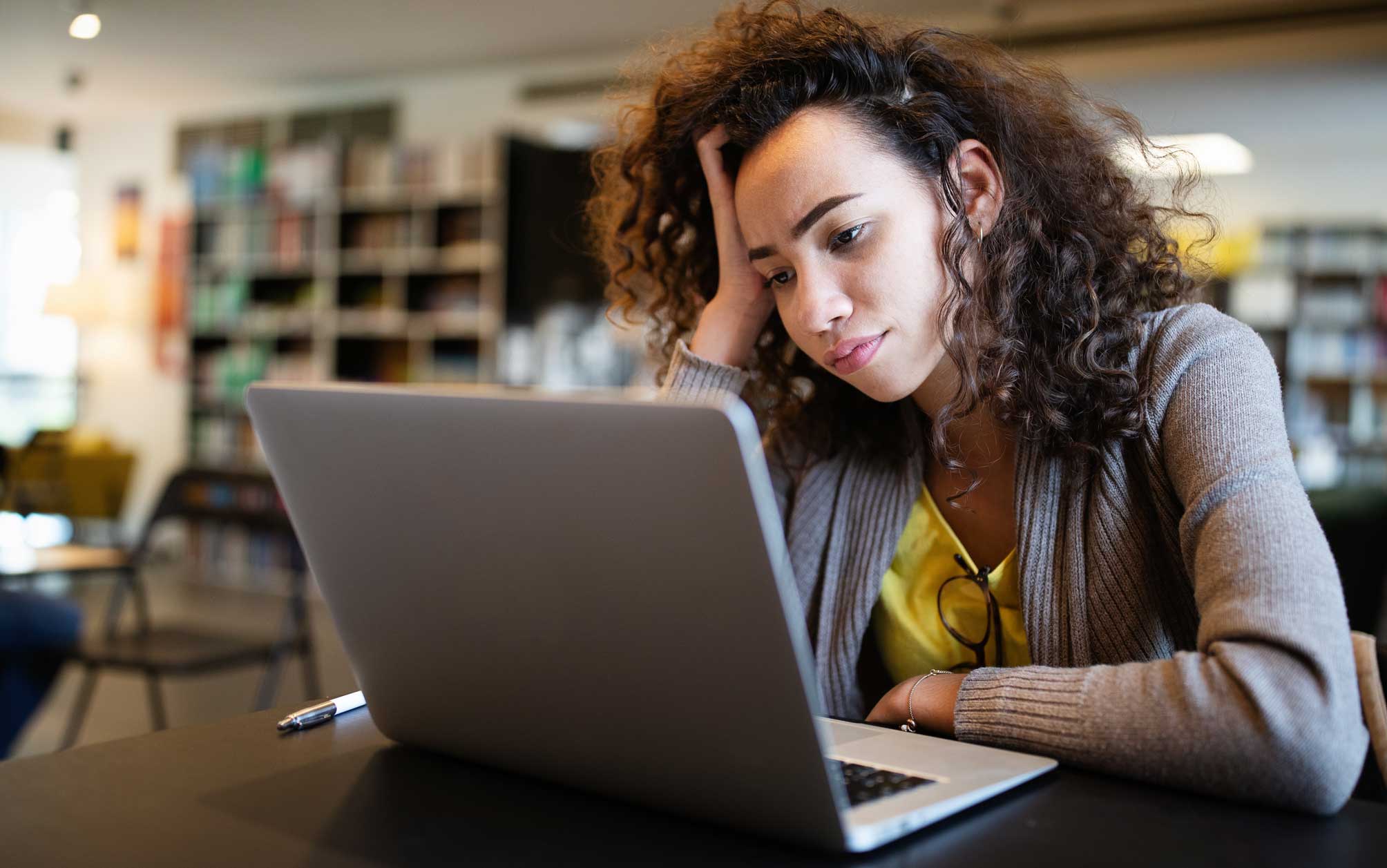 A young woman is looking confused at a laptop while working in a library.
