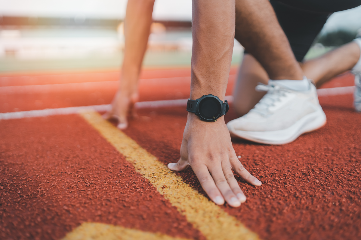 A close-up of a man’s hands and feet on a track oval as he is getting ready to start running. There is a black sports watch clearly visible on the man’s wrist.