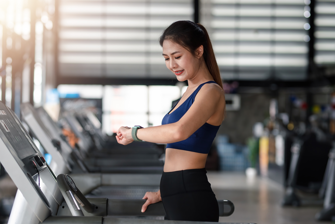 A young woman is standing on a treadmill at the gym. She is resting and looking at her sports watch.