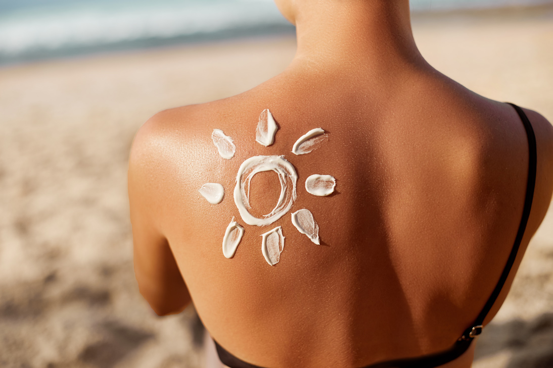 A woman is on a beach. The image shows her back only, and there is sunscreen on her left shoulder in the shape of a sun.