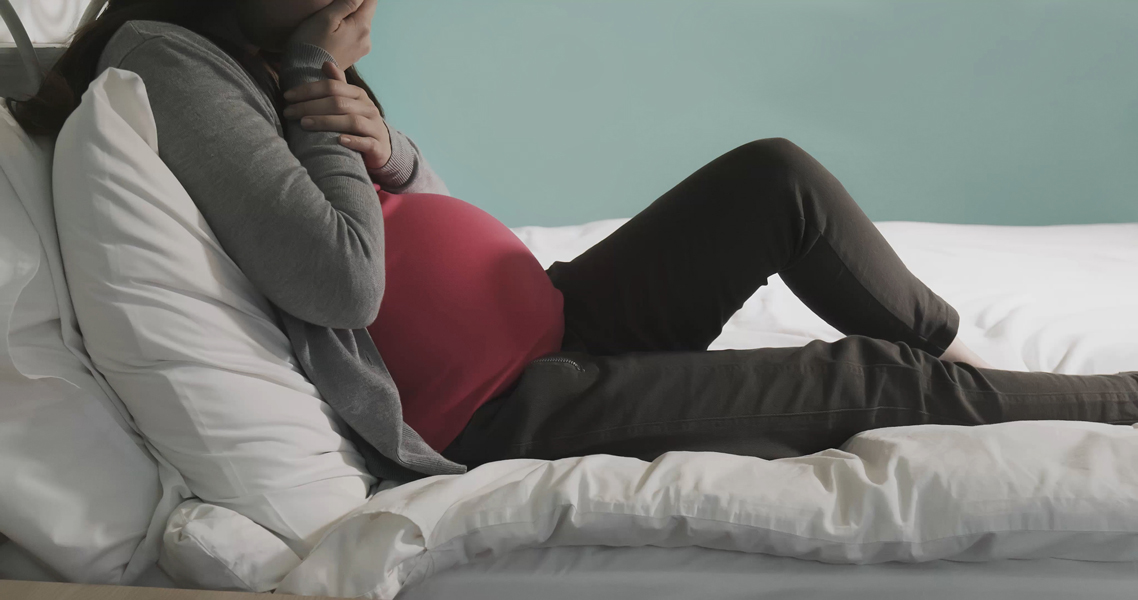 A pregnant woman on a bed is shown from the neck down. She is sitting up in the bed, and looks depressed, with one hand reaching up to her face.